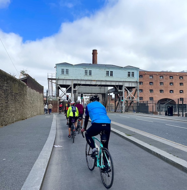 Cyclists going along the dock road. A blue bridge ahead with rooms.