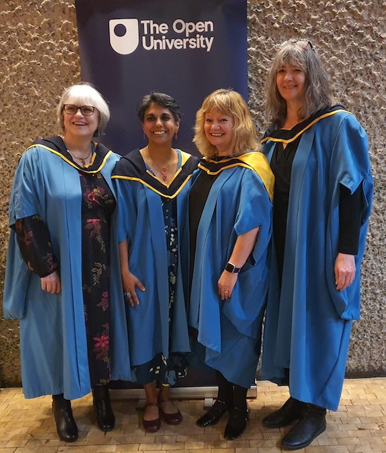 Four of us in our blue masters gowns in front of the Open University sign.