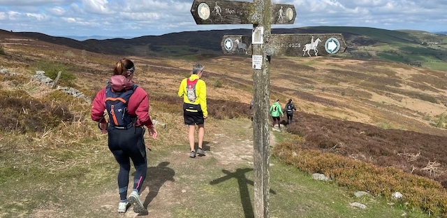Runners running along a path in the hills past a signpost